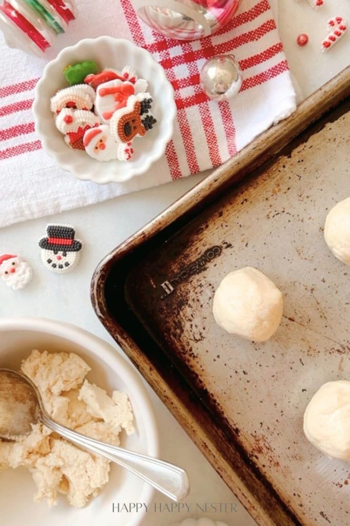 A baking sheet with unbaked Snow Globe Cookies, a bowl of raw dough with a spoon, and decorative holiday-themed candies rest on a white and red striped cloth. Snowman and Santa decorations are scattered around the scene, adding a festive touch to this cozy holiday preparation.