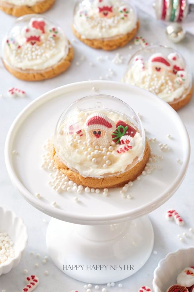 A festive cookie topped with white frosting, candy decorations, and white sprinkles is encased in a clear dome. It sits on a white pedestal. More similarly decorated cookies are in the background.