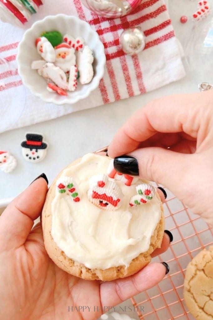 A hand meticulously decorates a frosted snow globe cookie with small holiday-themed sugar decorations. A red and white striped towel and a bowl brimming with more festive toppings are in the background on a table.