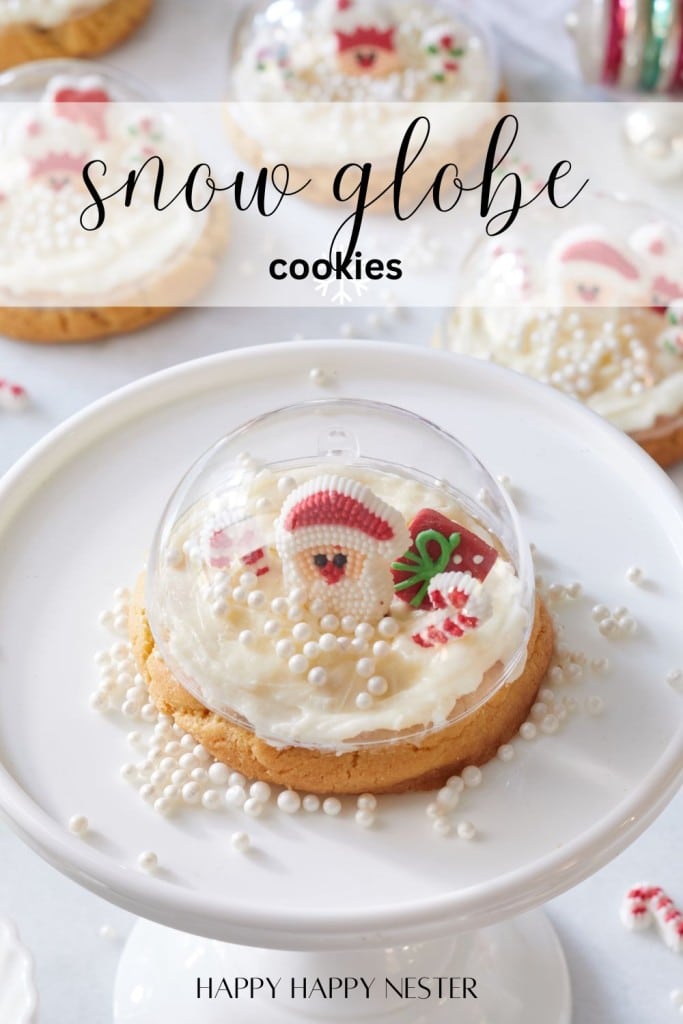 A festive cookie decorated with white frosting, sugar pearls, a small Santa face, and holiday-themed candies is encased in a clear plastic dome. The scene is set on a white dessert stand with the text "snow globe cookies" above.