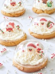 Five Snow Globe Cookies with frosting are topped with clear domes. Inside each dome, there are small Christmas-themed decorations, including Santa figures, candy canes, and snow-like white sprinkles, resembling magical holiday scenes.