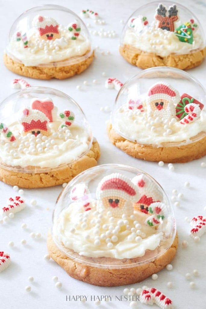 Five Snow Globe Cookies with frosting are topped with clear domes. Inside each dome, there are small Christmas-themed decorations, including Santa figures, candy canes, and snow-like white sprinkles, resembling magical holiday scenes.