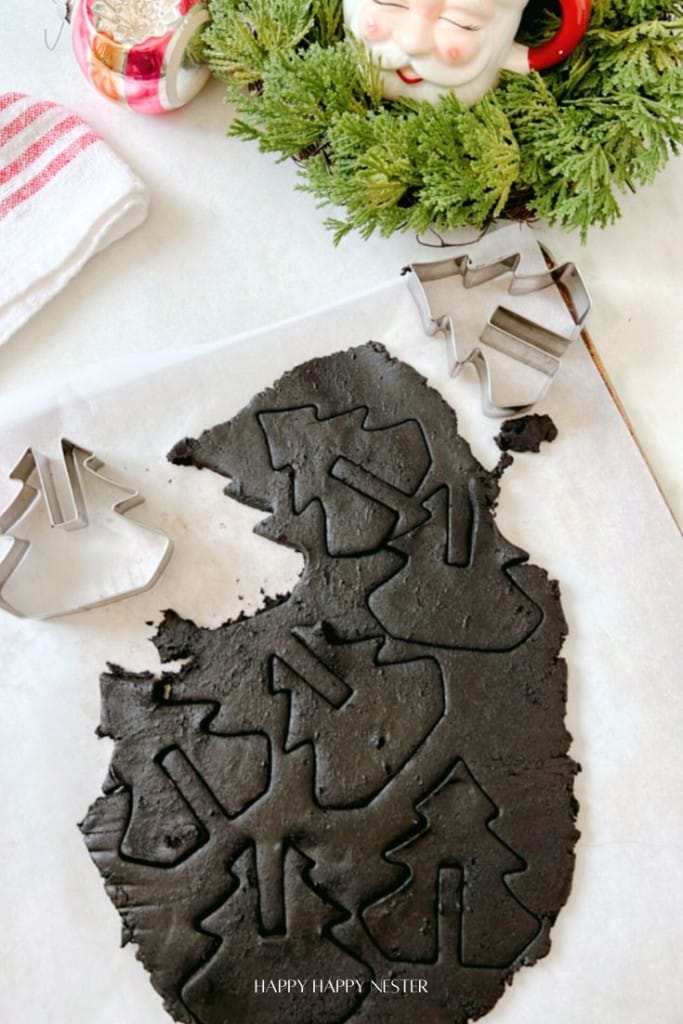 Christmas Tree Cookies take shape as cookie cutters press into dark dough on parchment paper. A Santa-themed mug and festive greenery complete the cozy holiday scene.