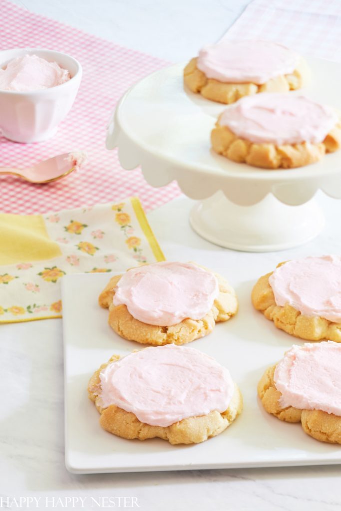 A plate of sugar cookies topped with light pink frosting sits on a white cake stand, embodying delightful New Year's recipes. More cookies are on a square white plate nearby. A pink-checkered napkin and floral cloth rest beneath a bowl with frosting and a spoon.