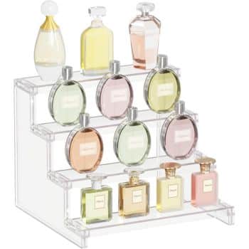 A clear acrylic display stand holds twelve perfume bottles of various shapes and colors on three tiers. The bottles contain liquids in shades of yellow, pink, and green, showcasing an elegant arrangement.