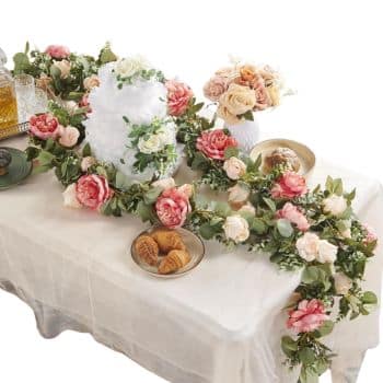 A decorated table with a white cake adorned with greenery and pink flowers. A garland of roses and eucalyptus runs along the table. A bouquet of roses in a white vase, pastries on a plate, and glass bottles are also present.