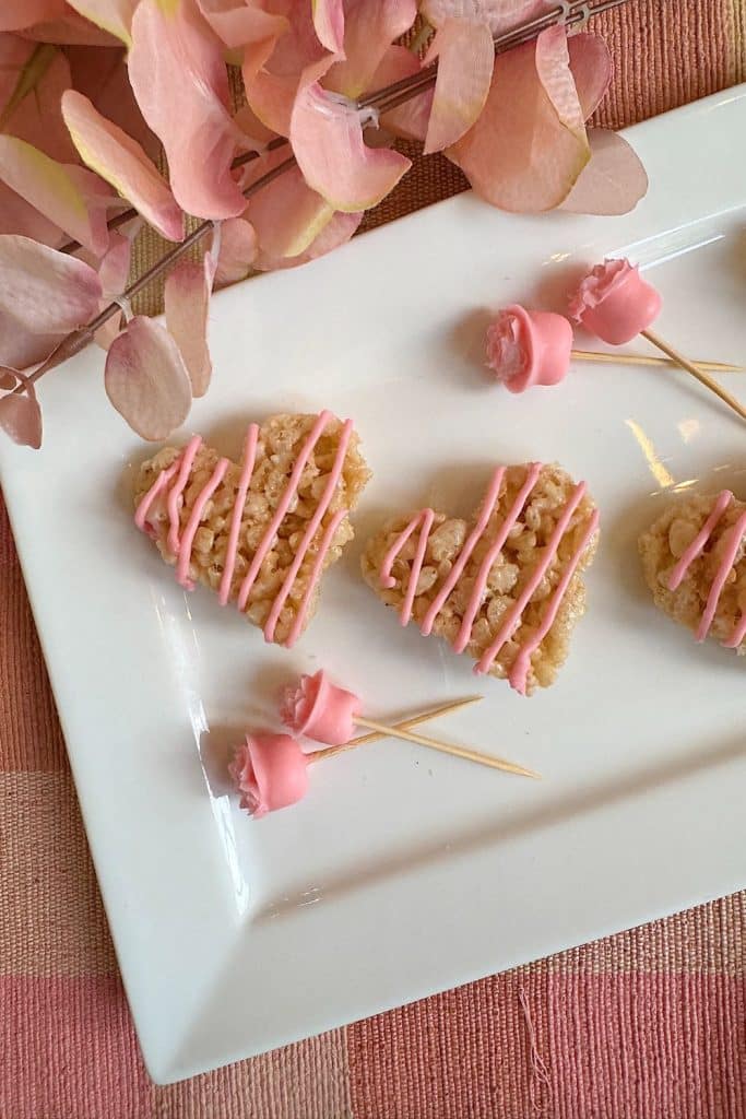 Heart-shaped rice treats drizzled with pink icing sit elegantly on a rectangular white plate, echoing the charm of an easy fruit trifle recipe. Accompanied by small pink rose-shaped decorations on wooden sticks, they rest on a pink checkered surface with pink leaves nearby.