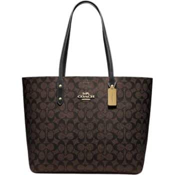 A brown Coach tote bag with a signature pattern and black leather straps. A small tag hangs from one strap, and the brand logo is displayed prominently on the front.