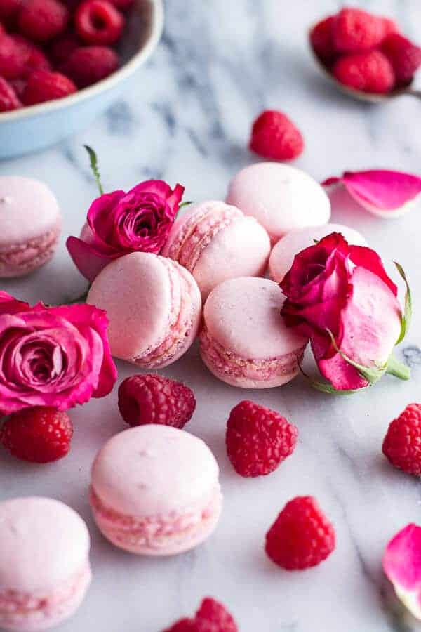 Pink macarons, delightful pink desserts, are surrounded by fresh raspberries and roses on a marble surface. A bowl of raspberries sits in the background, adding a touch of color and elegance to the composition.