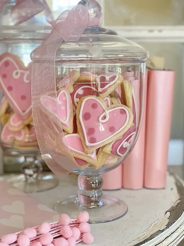 A glass jar filled with pink heart-shaped cookies, some adorned with polka dots and swirls, is topped with a pink ribbon. These delightful pink desserts are complemented by charming decor items, including cylindrical shapes and pom-poms.