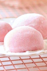 Close-up of Easy Mochi, pink desserts dusted with powdered sugar, resting on a metal cooling rack. The soft, round mochi have a smooth texture and gentle light pink color, evoking a sweet and delicate treat.