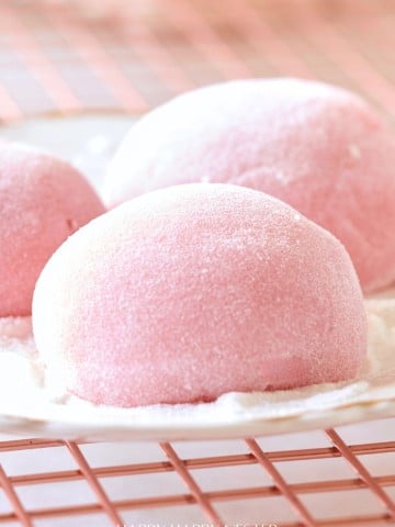 Close-up of Easy Mochi, pink desserts dusted with powdered sugar, resting on a metal cooling rack. The soft, round mochi have a smooth texture and gentle light pink color, evoking a sweet and delicate treat.