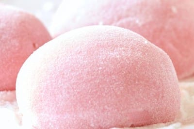 Close-up of Easy Mochi, pink desserts dusted with powdered sugar, resting on a metal cooling rack. The soft, round mochi have a smooth texture and gentle light pink color, evoking a sweet and delicate treat.