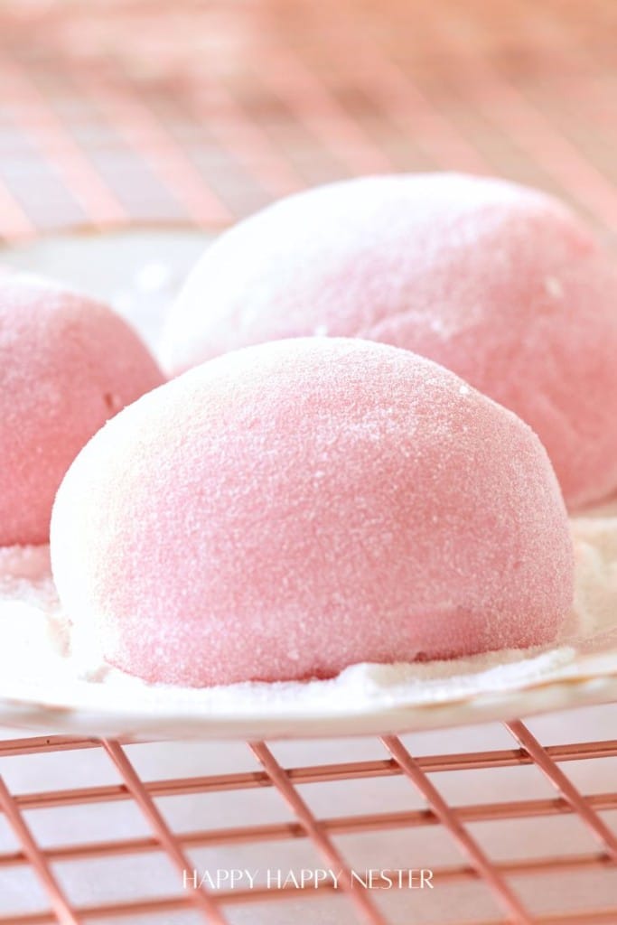 Close-up image of pink mochi ice cream balls on a white plate, placed on a grid-patterned pink surface. These delightful pink desserts are dusted with a light layer of powdered sugar, creating a soft and delicate appearance.