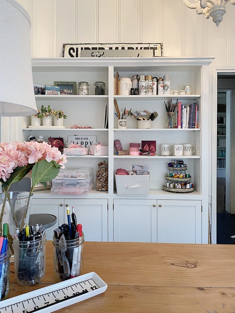 A cozy home office features a wooden table adorned with jars of pens and a vase of flowers. Behind it, a white bookshelf, organized with professional organizing tips, displays crafts and decorative items like mugs, books, and art supplies. A vintage "Super Market" sign adds charm on top.