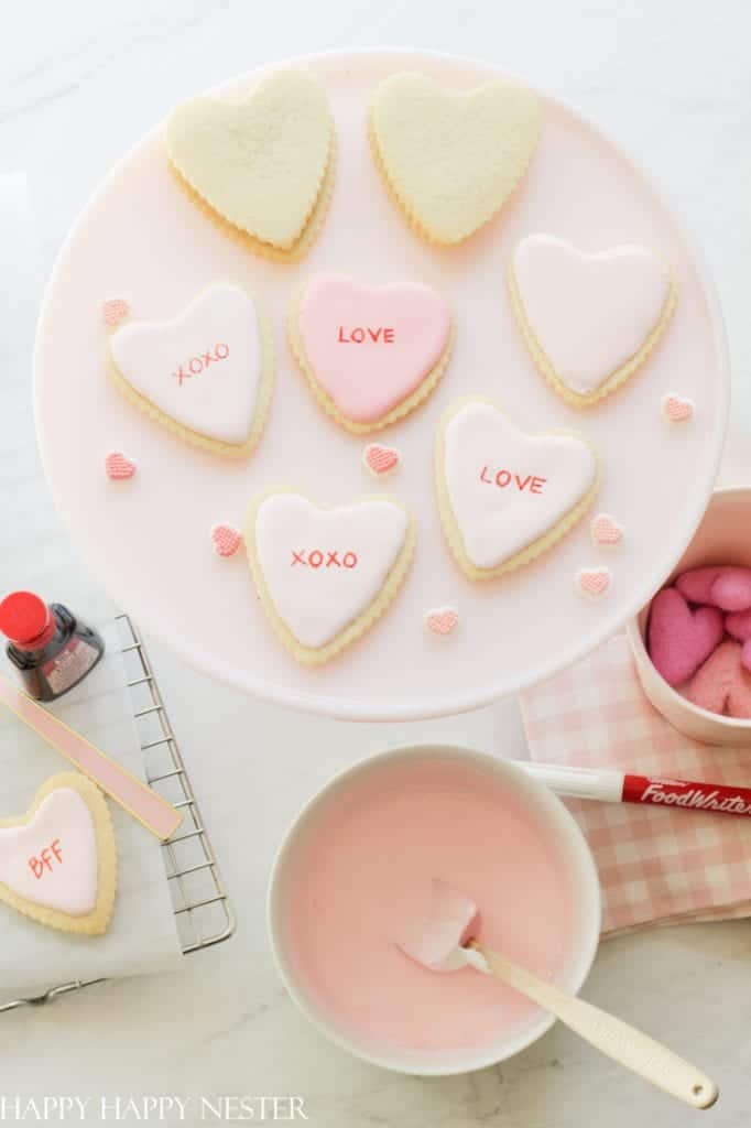 A delightful display of heart-shaped cookies on a white platter showcases pink desserts. Some feature pink icing with charming words like "LOVE," "XOXO," and "BFF." Nearby, a bowl of pink icing with a spatula, adorable pink heart candies, and a bottle of red food color complete the scene.