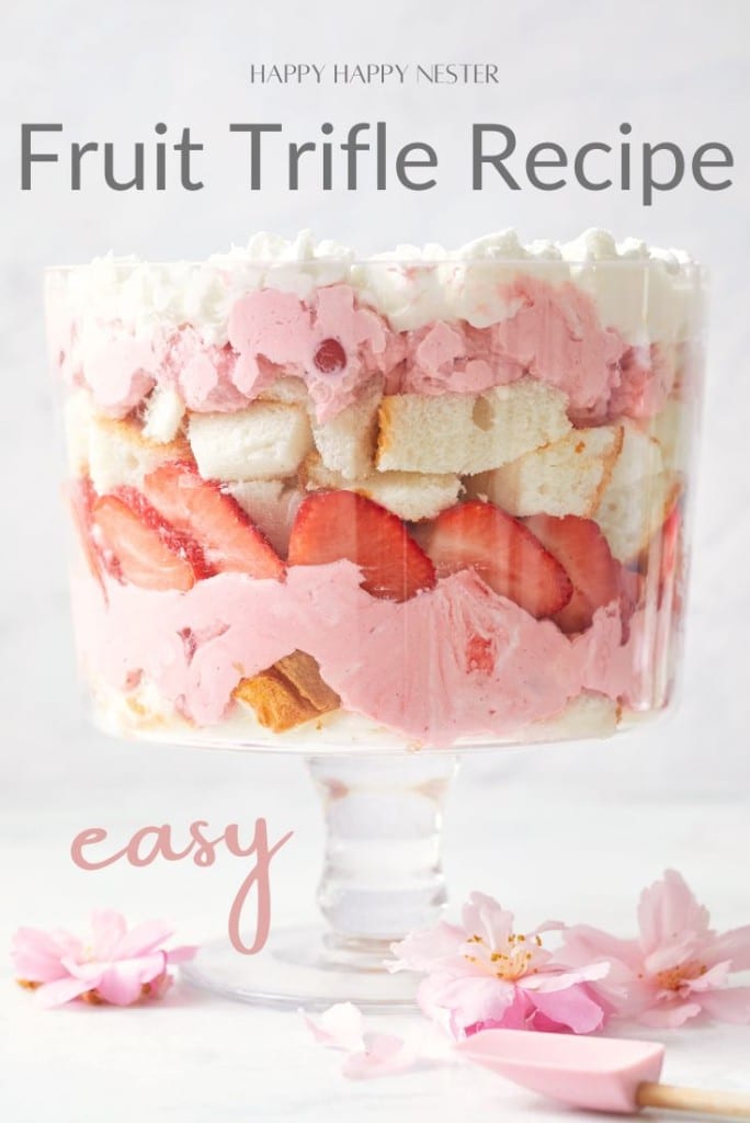 A layered fruit trifle in a glass bowl, showcasing strawberries, cake pieces, and pink whipped cream. The text reads "Happy Happy Nester Easy Fruit Trifle Recipe" and "easy." Pink flowers are scattered at the base.