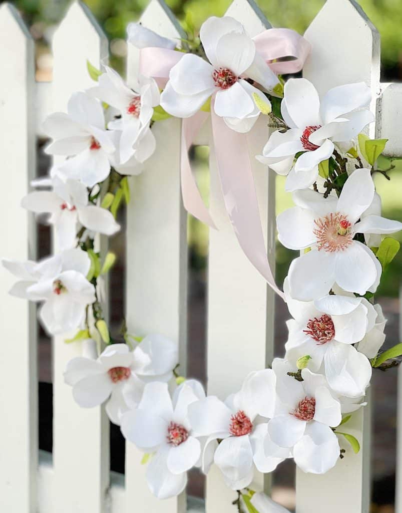 A white seasonal wreath with large, blooming flowers and pink centers hangs on a white picket fence. A pink ribbon is tied at the top, adding a decorative touch. The background features blurred greenery.