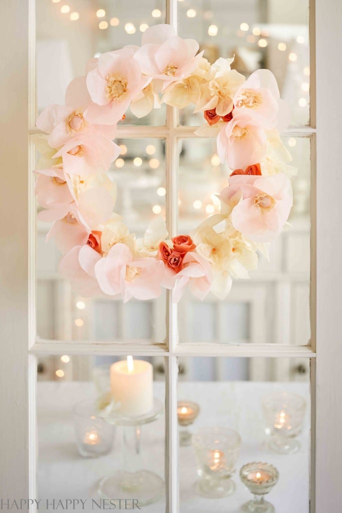 A floral wreath crafted with pink and white blooms adorns the glass door. Below, a lit candle is nestled among small votives, exuding a cozy ambiance. The soft lights blurred in the background enhance this pink crafts-inspired scene.