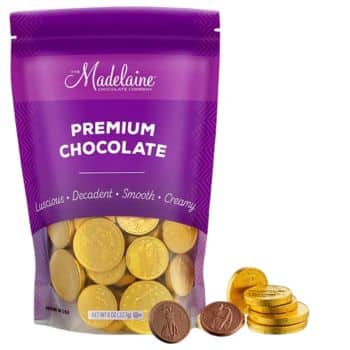 A purple bag labeled "Madelainé Premium Chocolate" stands upright. Nearby, gold foil-wrapped and unwrapped chocolate coins gleam enticingly. The bag boasts that these luscious, decadent treats are ideal not just for snacking but also for recipes made with chocolate chips—smooth and creamy perfection.