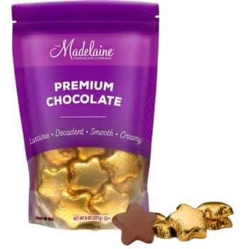 A purple bag labeled "The Madelaine Chocolate Company: Premium Chocolate" is shown. The bag contains gold-wrapped star-shaped chocolates, reminiscent of homemade recipes made with chocolate chips. Two chocolates are placed outside the bag, one unwrapped, revealing smooth milk chocolate.