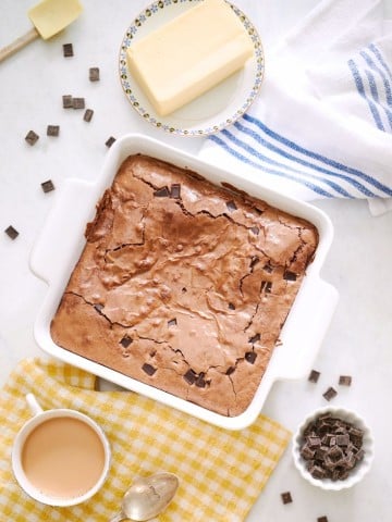 A white baking dish holds a batch of double chocolate brownies on a table adorned with butter, a steaming cup of coffee, a spoon, and rich chocolate chunks. A blue-striped towel rests nearby on a yellow checked cloth, completing this tempting scene.