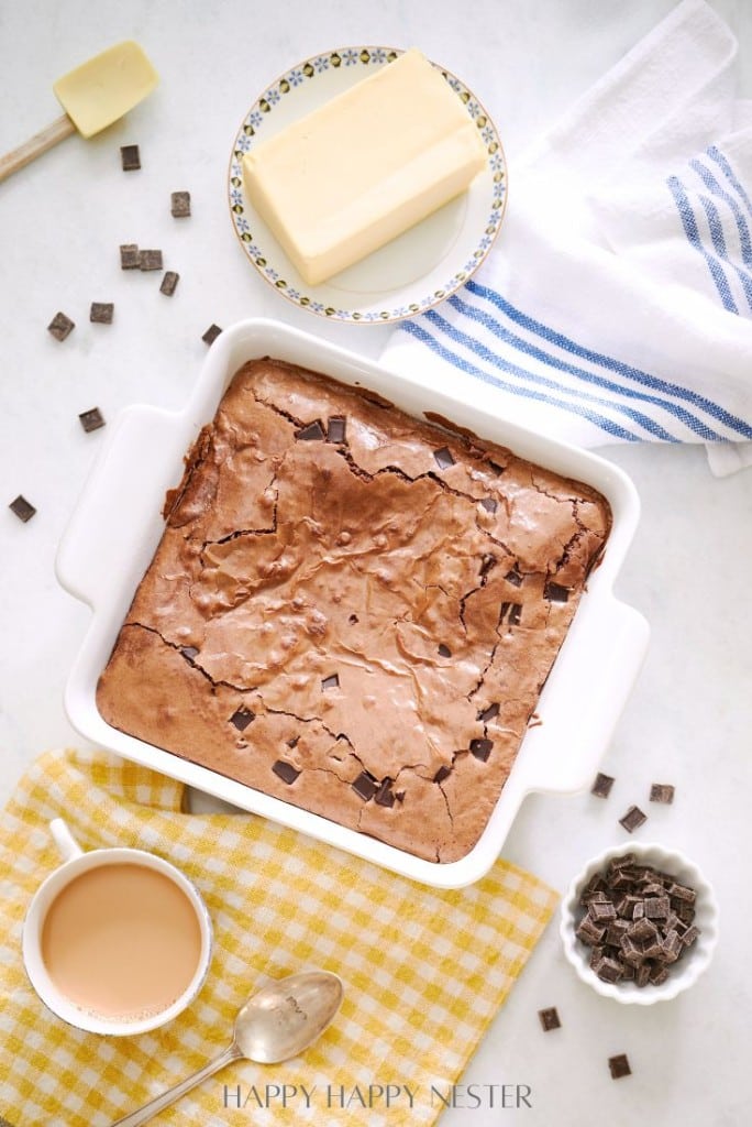 A white baking dish holds a batch of double chocolate brownies on a table adorned with butter, a steaming cup of coffee, a spoon, and rich chocolate chunks. A blue-striped towel rests nearby on a yellow checked cloth, completing this tempting scene.