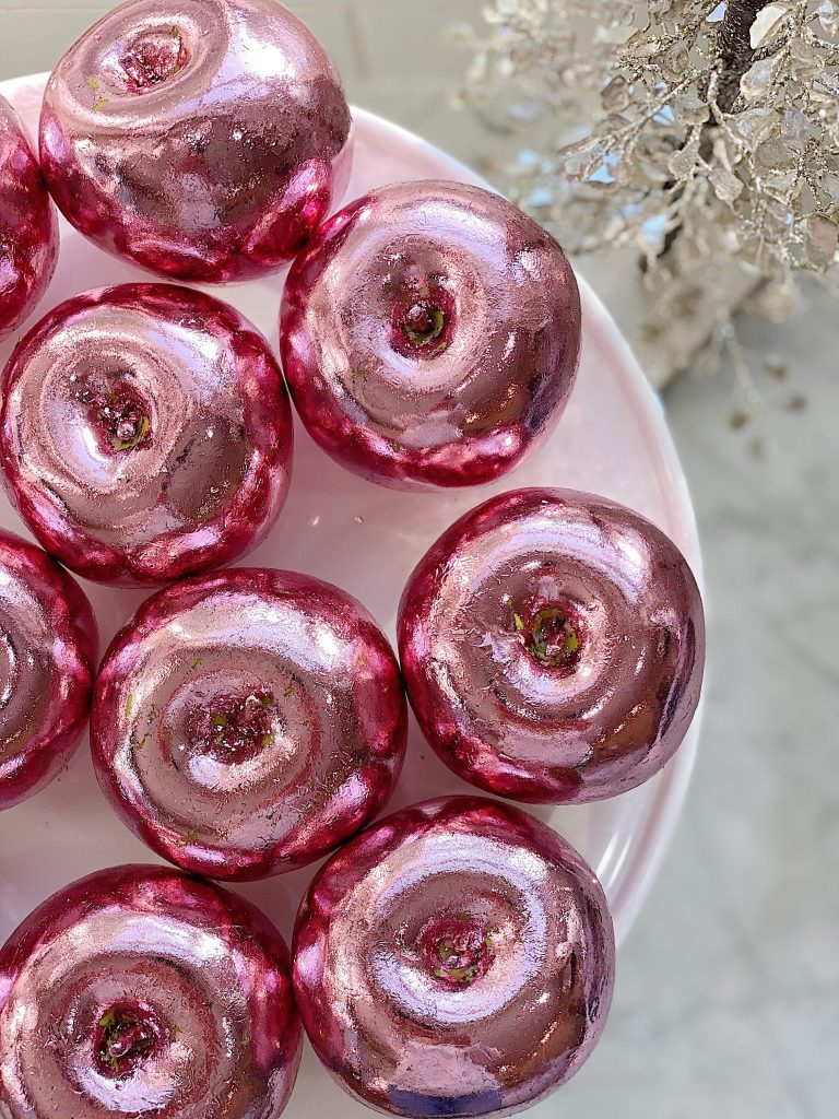 A plate of shiny, metallic pink glazed donuts arranged in a circle, reminiscent of intricate pink crafts, displayed on a white surface. In the background, a decorative, glittery silver plant adds a festive touch to the scene.