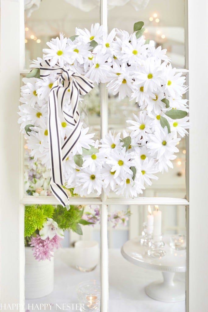 A white daisy seasonal wreath with a black and white striped ribbon hangs on a glass window. Behind it, a table with flowers, lit candles on a silver tray, and a blurred background of white lights is visible.