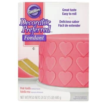 Package of Wilton Decorator Preferred Fondant in pink vanilla flavor, perfect for crafting delightful pink desserts. Features a pink cake adorned with heart shapes. Text highlights "Great taste, easy to roll" in English and Spanish. Net weight: 24 oz (680 g).