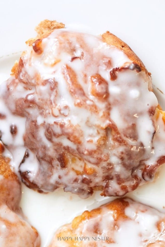 A close-up of a glazed cinnamon roll with creamy white icing covering most of its golden-brown surface, slightly melting and glistening under the light, reminiscent of the luscious texture in a classic apple fritter recipe.