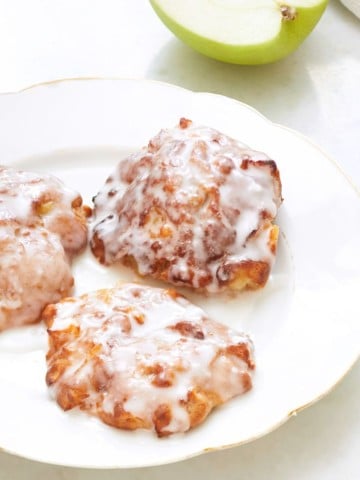 Three apple fritters with a white glaze are on a white plate. A green apple is partially visible in the background.