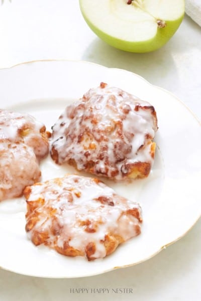 Three apple fritters with a white glaze are on a white plate. A green apple is partially visible in the background.