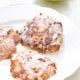 Three apple fritters with a white glaze are on a white plate. A green apple is partially visible in the background.