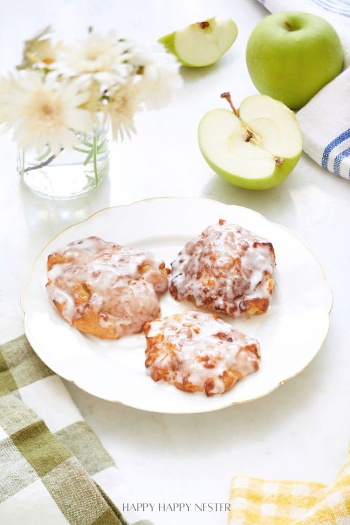 A white plate with three glazed apple fritters is on a table. Nearby are green apples, a vase with white flowers, and colorful checkered cloth napkins. The scene is bright and inviting, suggesting a cozy kitchen setting.