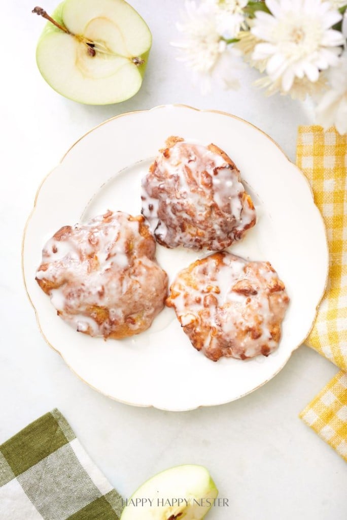 Three glazed apple fritters rest on a white plate, embodying the perfect baked apple fritter recipe. A sliced green apple, white flowers, and checkered cloths in green and yellow surround the dish, all set against a light, textured background.