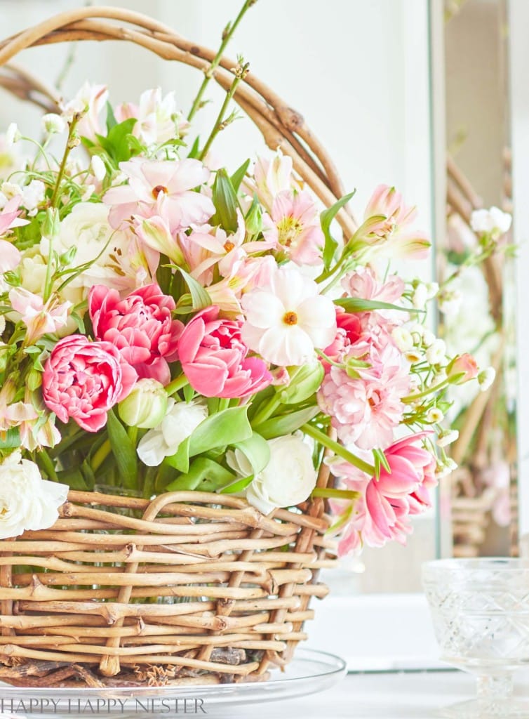 A wicker basket brimming with a variety of fresh flowers, including pink tulips and white blossoms, sits on a glass surface. Green leaves weave through the blooms like an artist's touch on pink crafts, creating a vibrant and colorful arrangement.