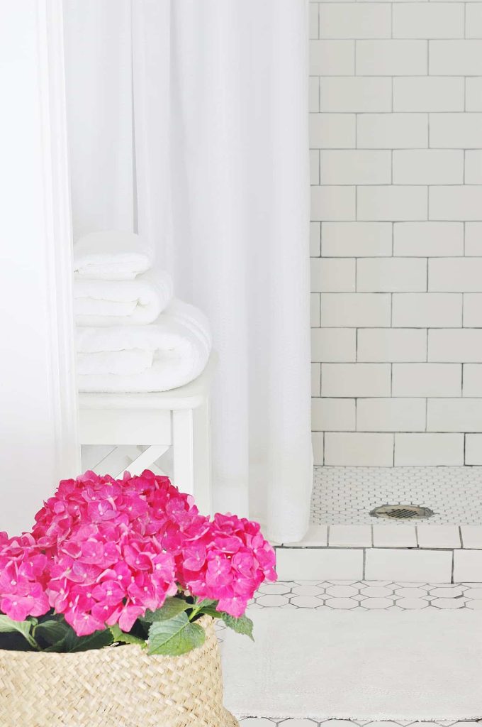 A bathroom with white tiled walls and floor. A shower area is partially visible behind a white curtain. For professional organizing tips, note the stack of white towels on a stool and a woven basket holding vibrant pink flowers in the foreground, adding both function and flair.