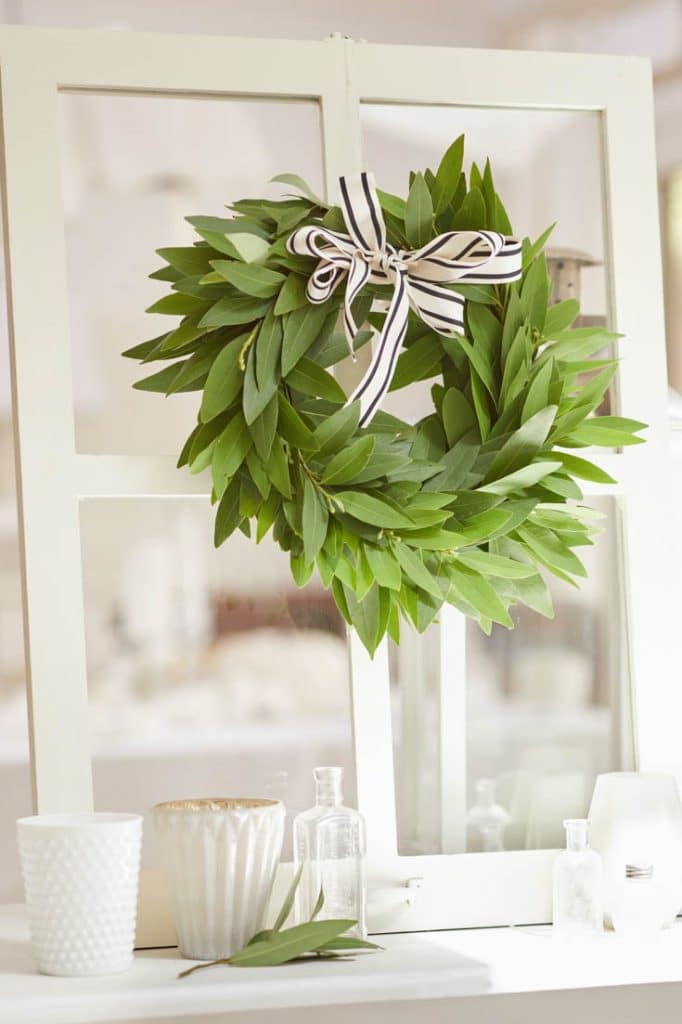 A seasonal wreath adorned with a black and white striped bow graces a white-framed window. Below, two white cups, glass bottles, and a small branch rest on a pristine surface, while the background remains softly blurred.