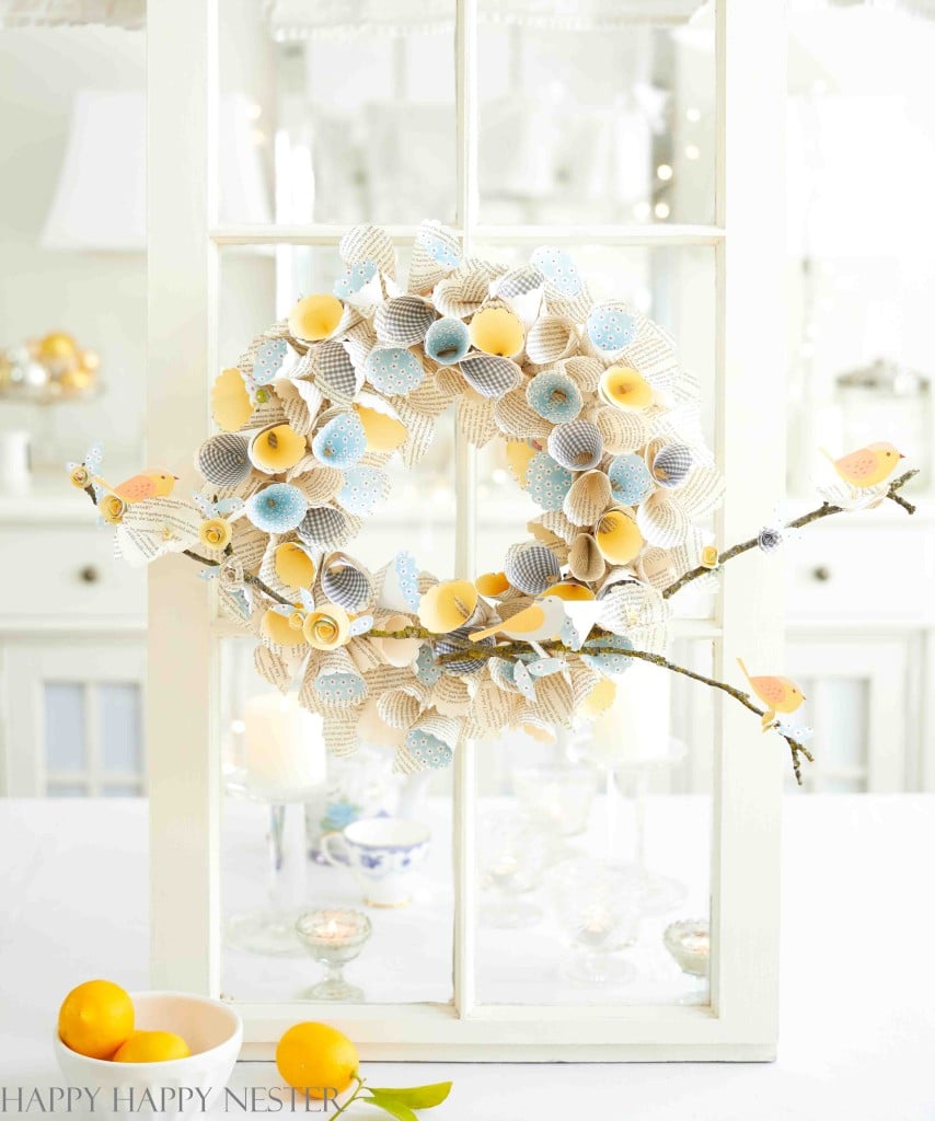 A delicate seasonal wreath made of paper flowers in soft yellow and light blue hues hangs on a white-framed glass door. Below, a small branch with paper bird decorations adds charm. A few lemons rest on the white table beneath, completing this picturesque scene.