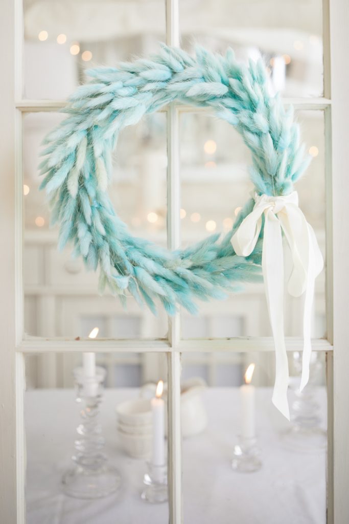 A fluffy, light blue seasonal wreath with a white ribbon hangs on a glass door. In the background, lit white candles on a table create a cozy and festive atmosphere.