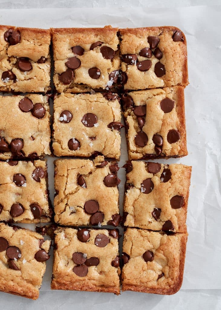 A batch of chocolate chip blondies cut into 12 squares rests on parchment paper. These golden-brown delights, perfect examples of recipes made with chocolate chips, boast melted morsels scattered across the top.