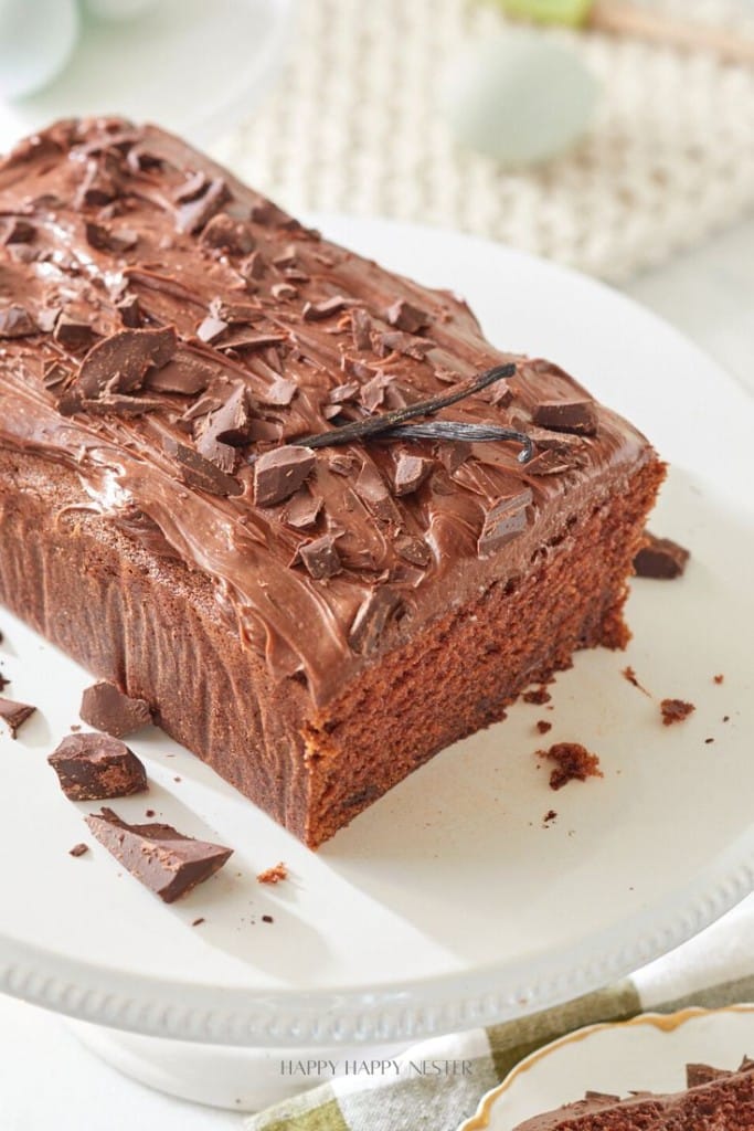 A chocolate loaf cake on a white cake stand. The cake is topped with chocolate frosting and garnished with chopped chocolate pieces. A slice has been cut from one end, revealing the moist interior.