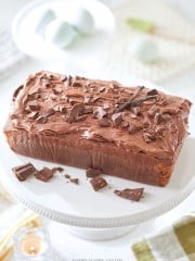 A loaf cake topped with chocolate frosting and chocolate shavings sits on a white cake stand. A vanilla bean pod is placed on top for decoration. The background includes a candle and blurred objects, creating a warm, inviting atmosphere.