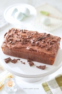 A loaf cake topped with chocolate frosting and chocolate shavings sits on a white cake stand. A vanilla bean pod is placed on top for decoration. The background includes a candle and blurred objects, creating a warm, inviting atmosphere.