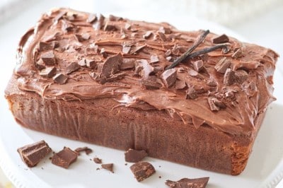 A loaf cake topped with chocolate frosting and chocolate shavings sits on a white cake stand. A vanilla bean pod is placed on top for decoration. The background includes a candle and blurred objects, creating a warm, inviting atmosphere.