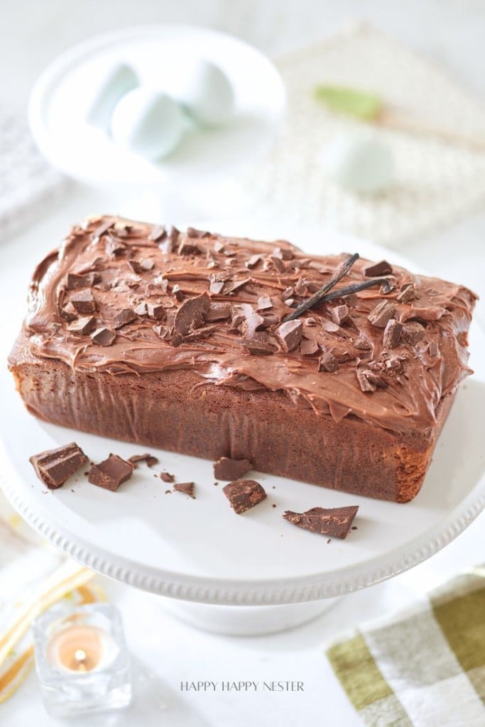 A loaf cake topped with chocolate frosting and chocolate shavings sits on a white cake stand. A vanilla bean pod is placed on top for decoration. The background includes a candle and blurred objects, creating a warm, inviting atmosphere.