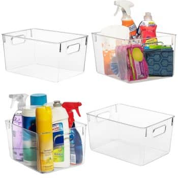 Four clear plastic storage bins are shown, perfect for implementing professional organizing tips. Two are empty, while the others neatly hold various cleaning supplies like spray bottles, detergent, and sponges. Each bin features side handles for easy carrying and effortless organization.