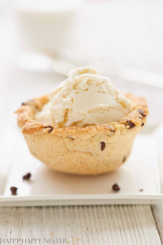 A scoop of vanilla ice cream rests atop a chocolate chip cookie cup, showcasing one of the delightful recipes made with chocolate chips. It's presented on a white plate, while the softly blurred background highlights its tempting allure.