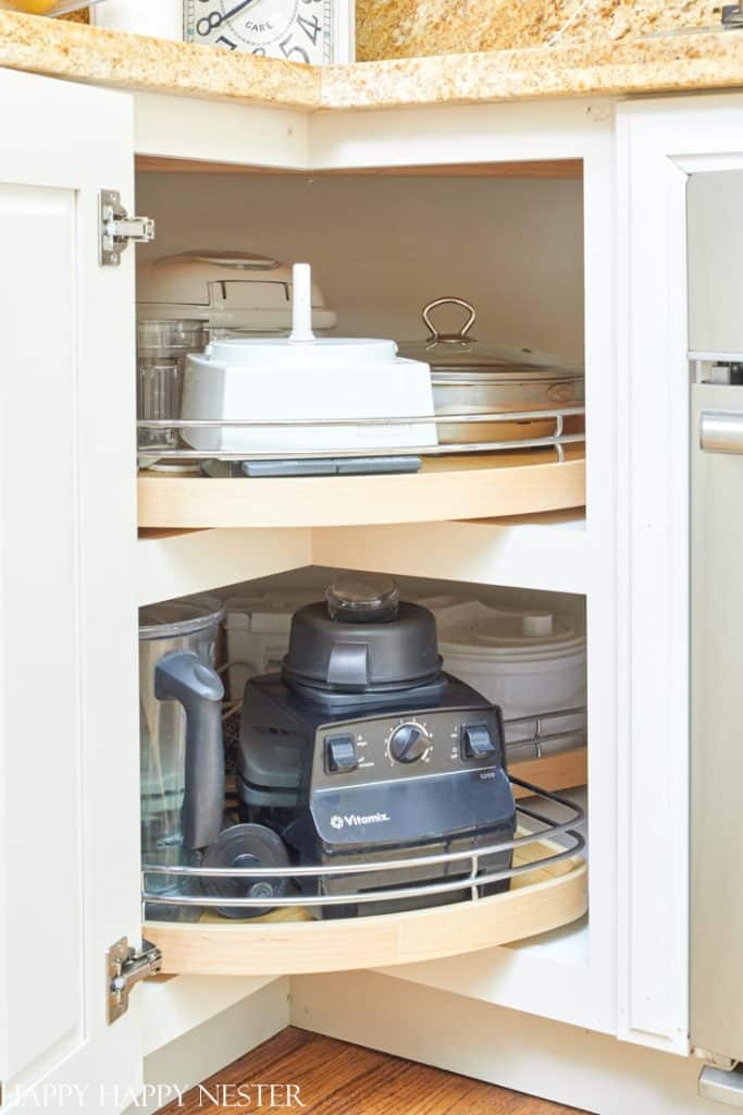 Corner kitchen cabinet with two lazy Susan shelves. Top shelf holds a cake carrier and metal trays, while the bottom shelf contains a Vitamix blender and food processor. Following professional organizing tips, wooden cabinet doors with silver handles open to reveal perfectly arranged storage.